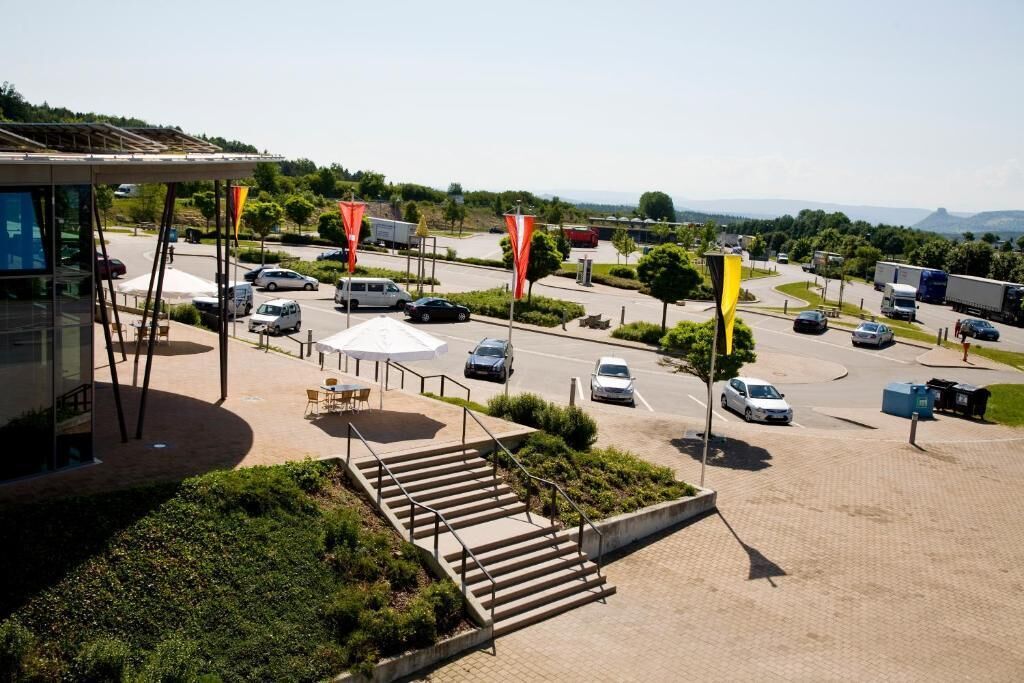 Un espace de stationnement avec plusieurs voitures, un espace de repos couvert et un bâtiment doté d’une façade en verre.