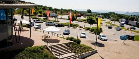 Un espace de stationnement avec plusieurs voitures, un espace de repos couvert et un bâtiment doté d’une façade en verre.