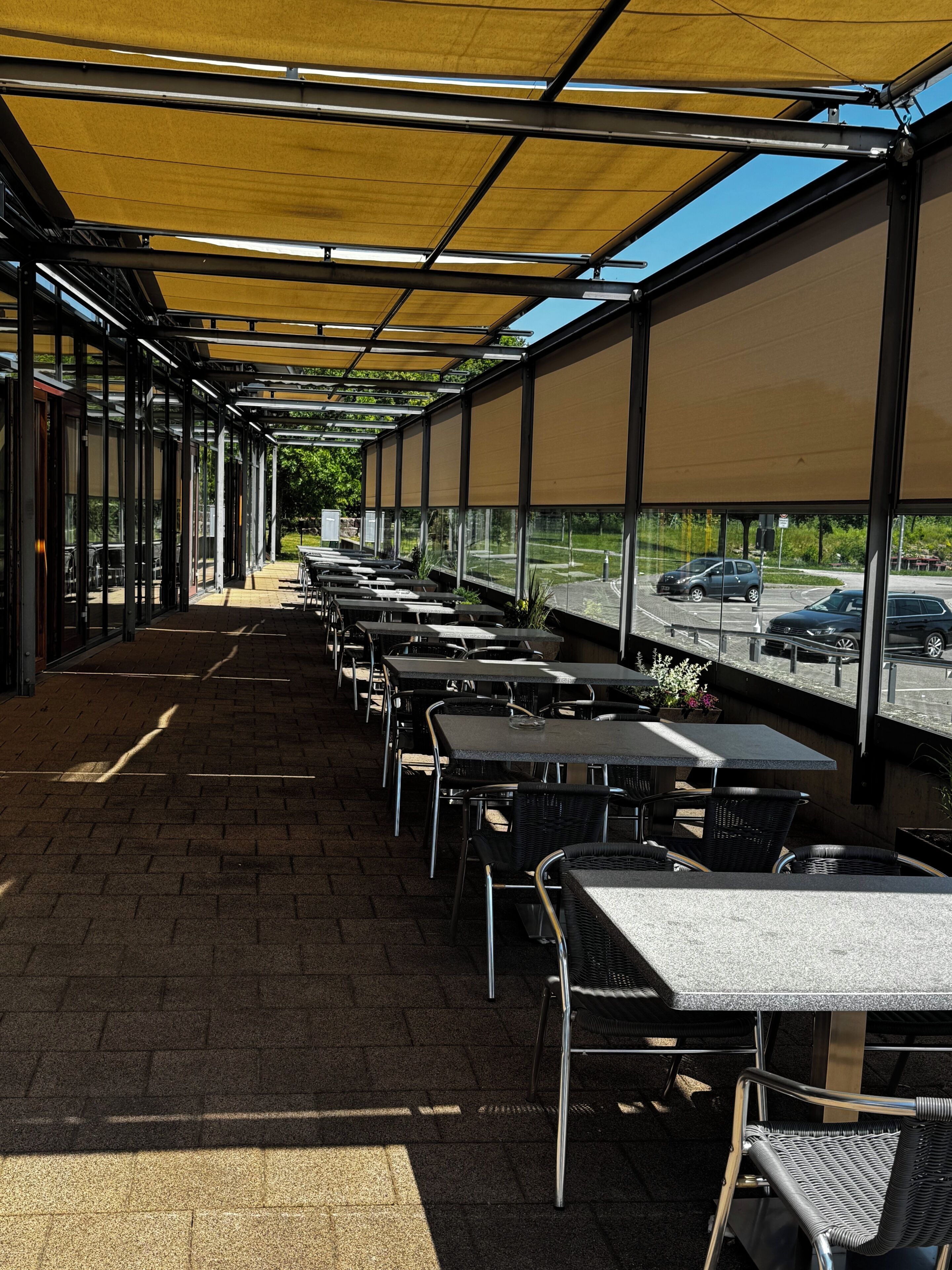 Un'area salotto esterna coperta con tavoli e sedie, un vialetto lastricato e vista su un parcheggio attraverso ampie finestre.