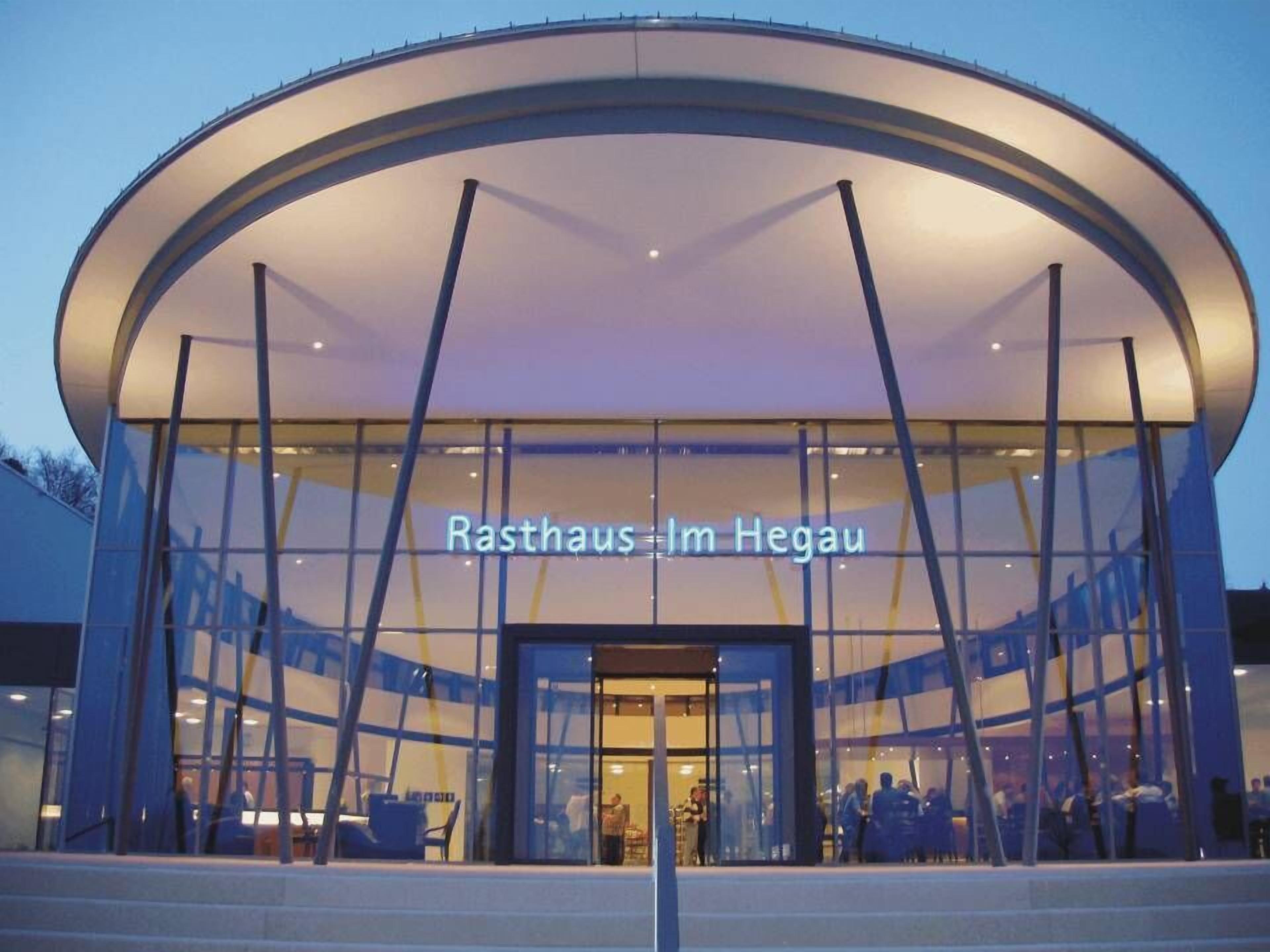 En moderne bygning med en buet glasfacade og teksten 'Rasthaus Im Hegau', der angiver et hostel eller indkvartering.