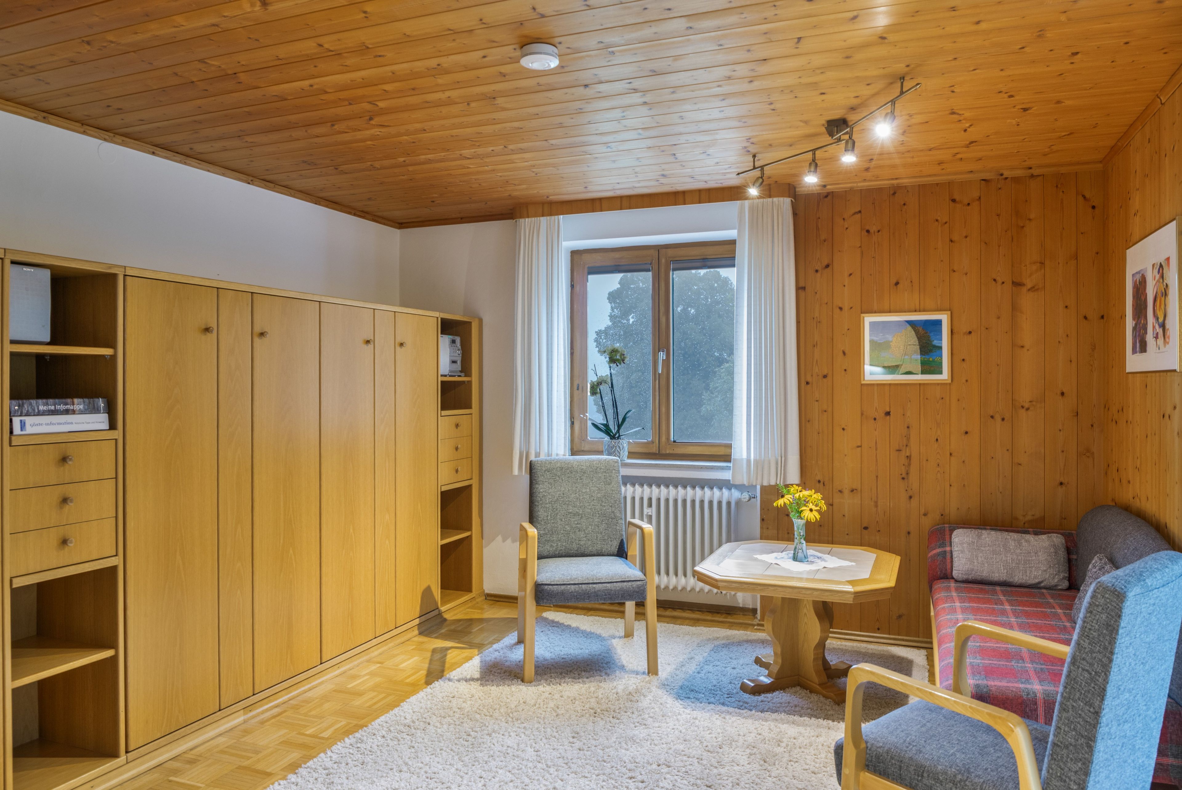A room with wooden walls and ceiling, a window with curtains, a radiator, a small table with a vase, a sofa, and a wardrobe with open doors.