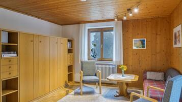 A room with wooden walls and ceiling, a window with curtains, a radiator, a small table with a vase, a sofa, and a wardrobe with open doors.