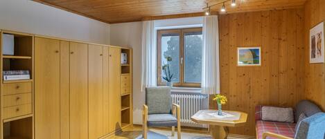 A room with wooden walls and ceiling, a window with curtains, a radiator, a small table with a vase, a sofa, and a wardrobe with open doors.