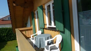 Ein Holzbalkon mit Tisch und Stühlen, ein Fenster mit Rollläden und eine Tür.