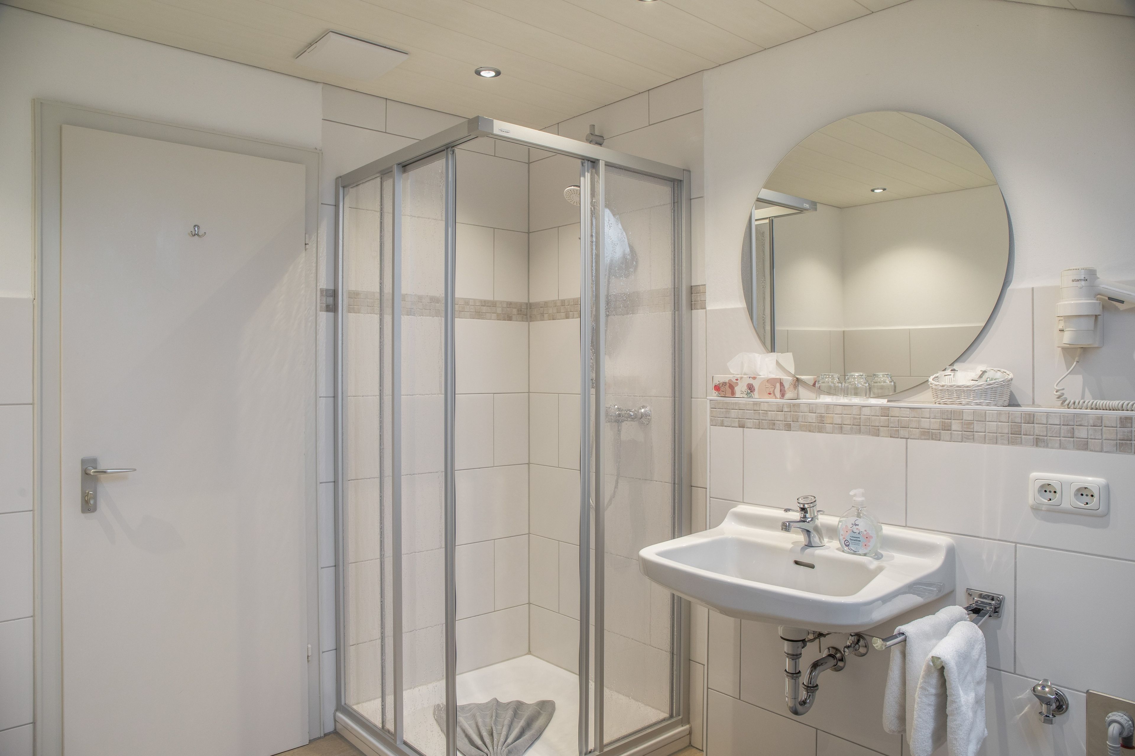 Et moderne badeværelse med brusekabine i glas, et håndvaskarrangement med spejl og toiletartikler på bordet.