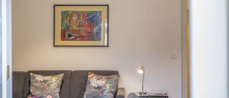 A living room with a grey sofa, a framed abstract painting, a small table with a vase of flowers, and a lamp on a side table.