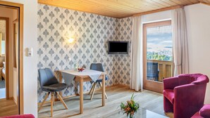 Een kamer met een houten plafond, een eettafel met stoelen, een aan de muur bevestigde televisie en een raam met gordijnen.