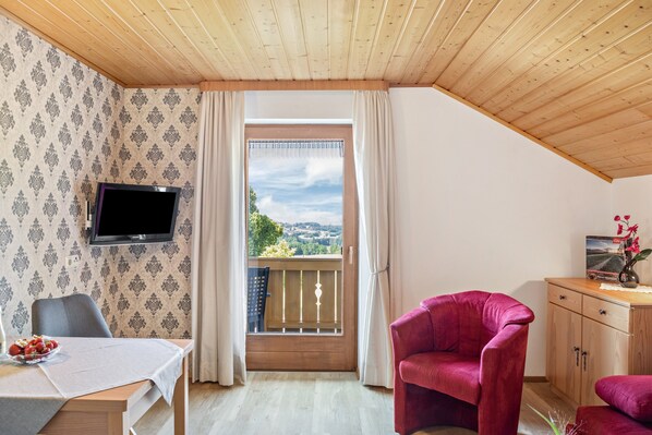 A room with a wooden ceiling, a TV mounted on the wall, a balcony with a view, a table with a placemat, and a red chair.