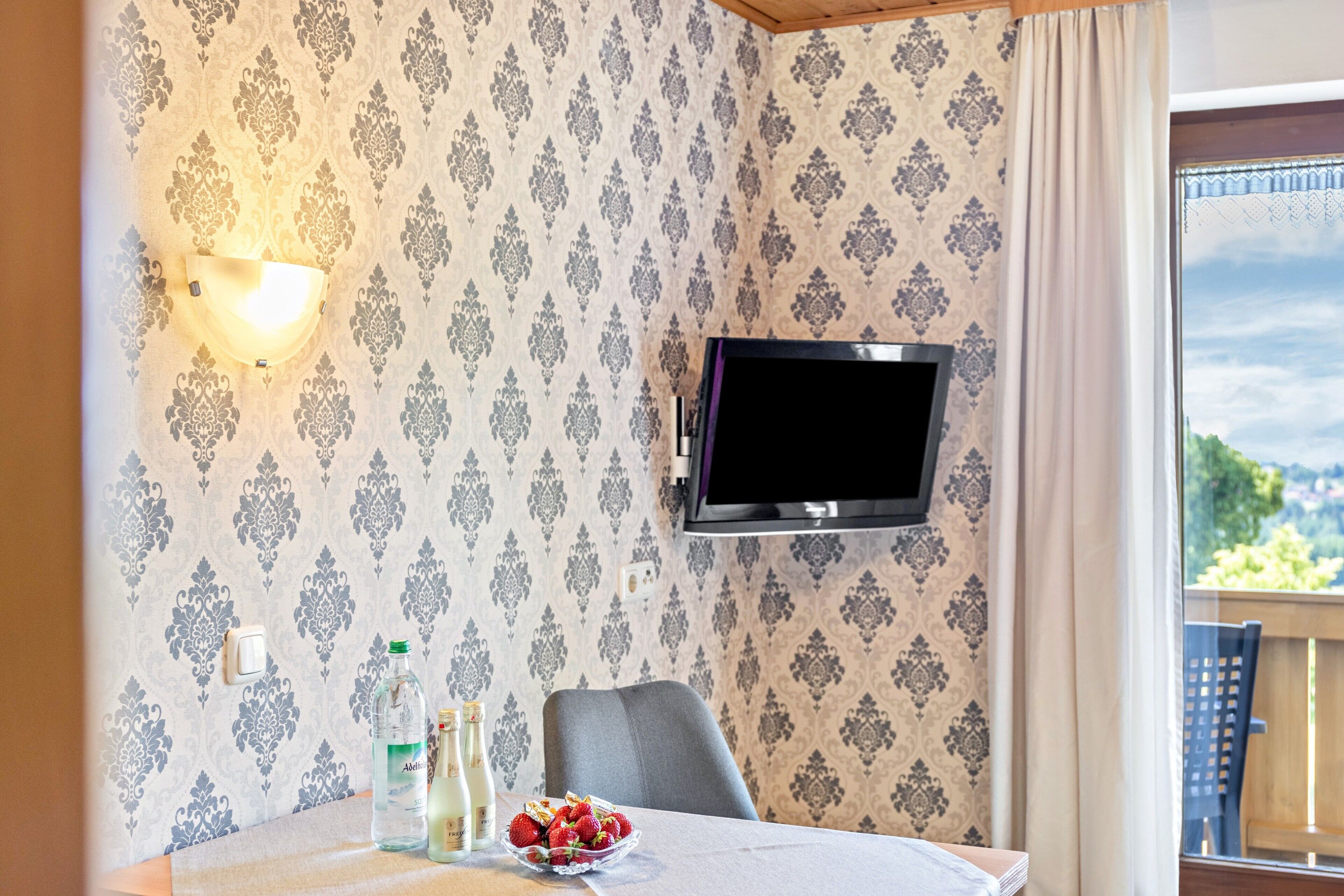 A room with patterned wallpaper, a flat-screen TV, a table with a bottle, a bowl of fruit, and a chair.