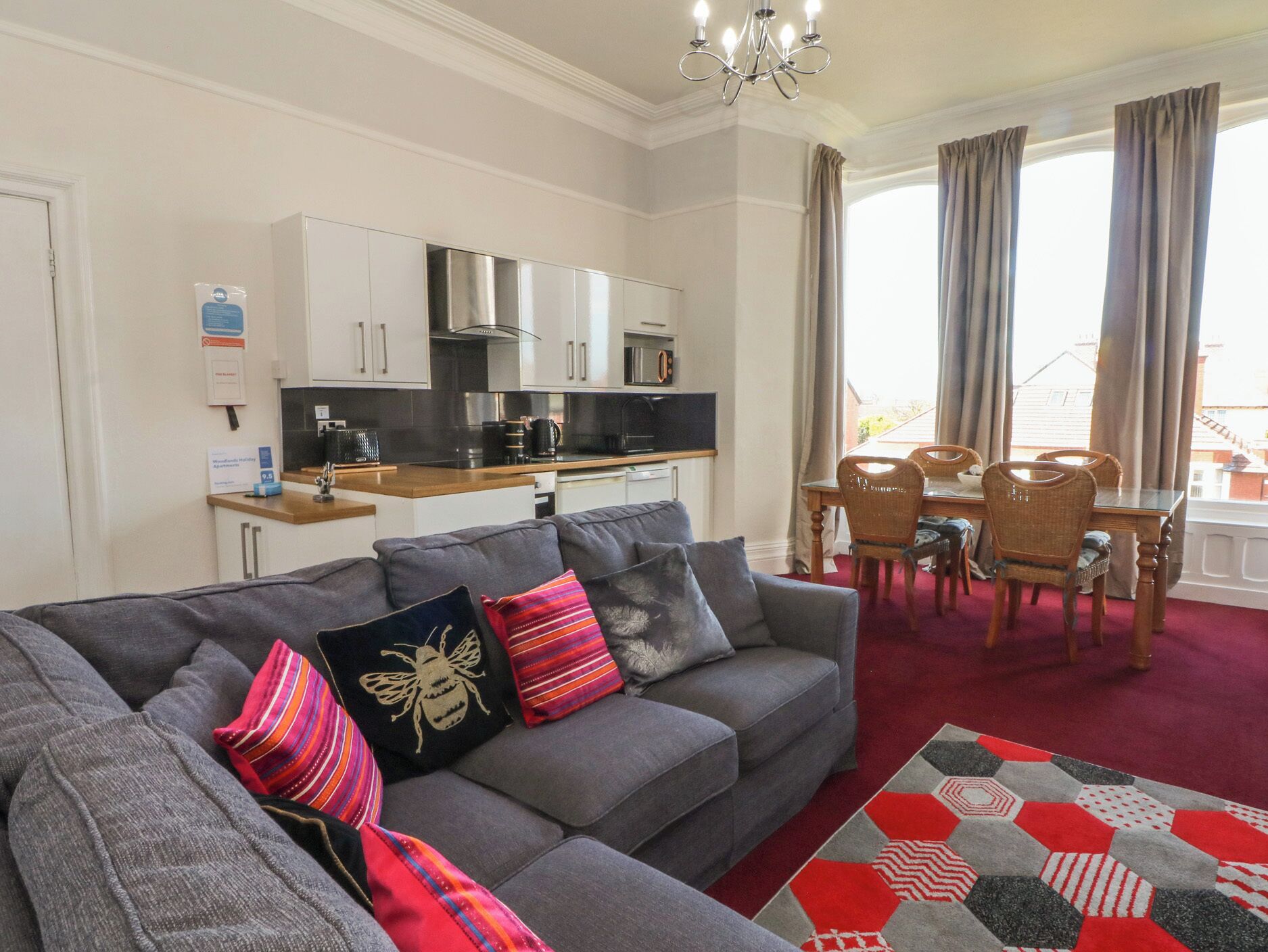A modern living room with a grey sofa, a dining area with a wooden table and chairs, and a kitchen with white cabinets and a stainless steel range hood.