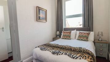 A hotel room with a bed, a nightstand, a lamp, a window with curtains, a framed picture on the wall, and a door.