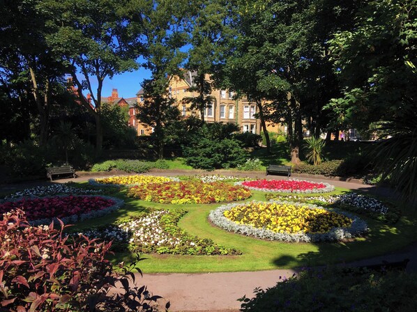 A garden with flower beds and benches, surrounded by trees and a building in the background. - Apartment 2 (Lytham St. Anne's)