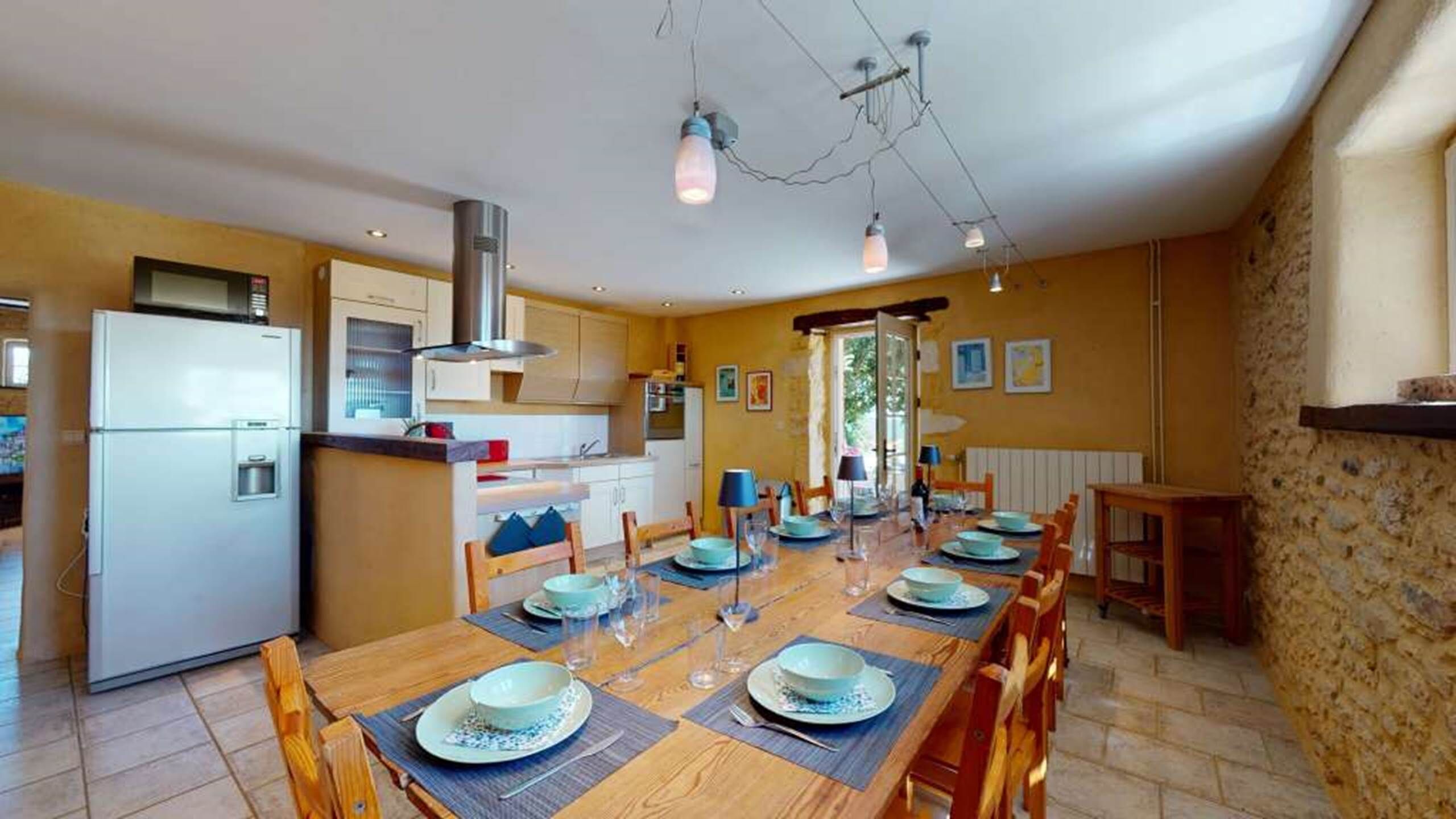 Un espace repas avec une longue table en bois pouvant accueillir six personnes, une cuisine moderne dotée d’un réfrigérateur intégré et d’un éclairage suspendu.