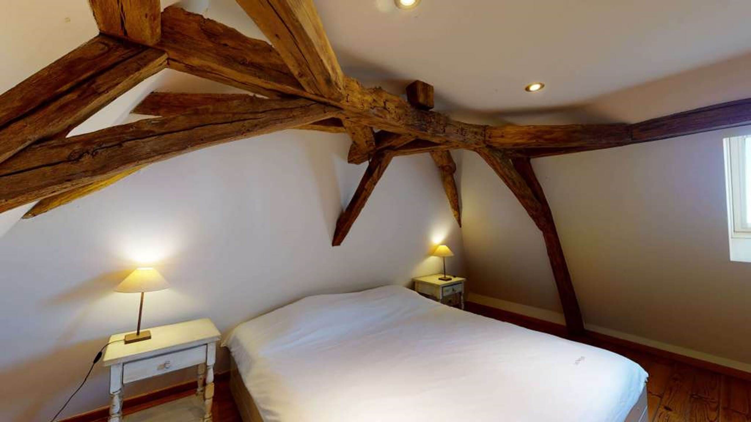 A bedroom with a sloped ceiling, exposed wooden beams, a bed, bedside tables, and two lamps.