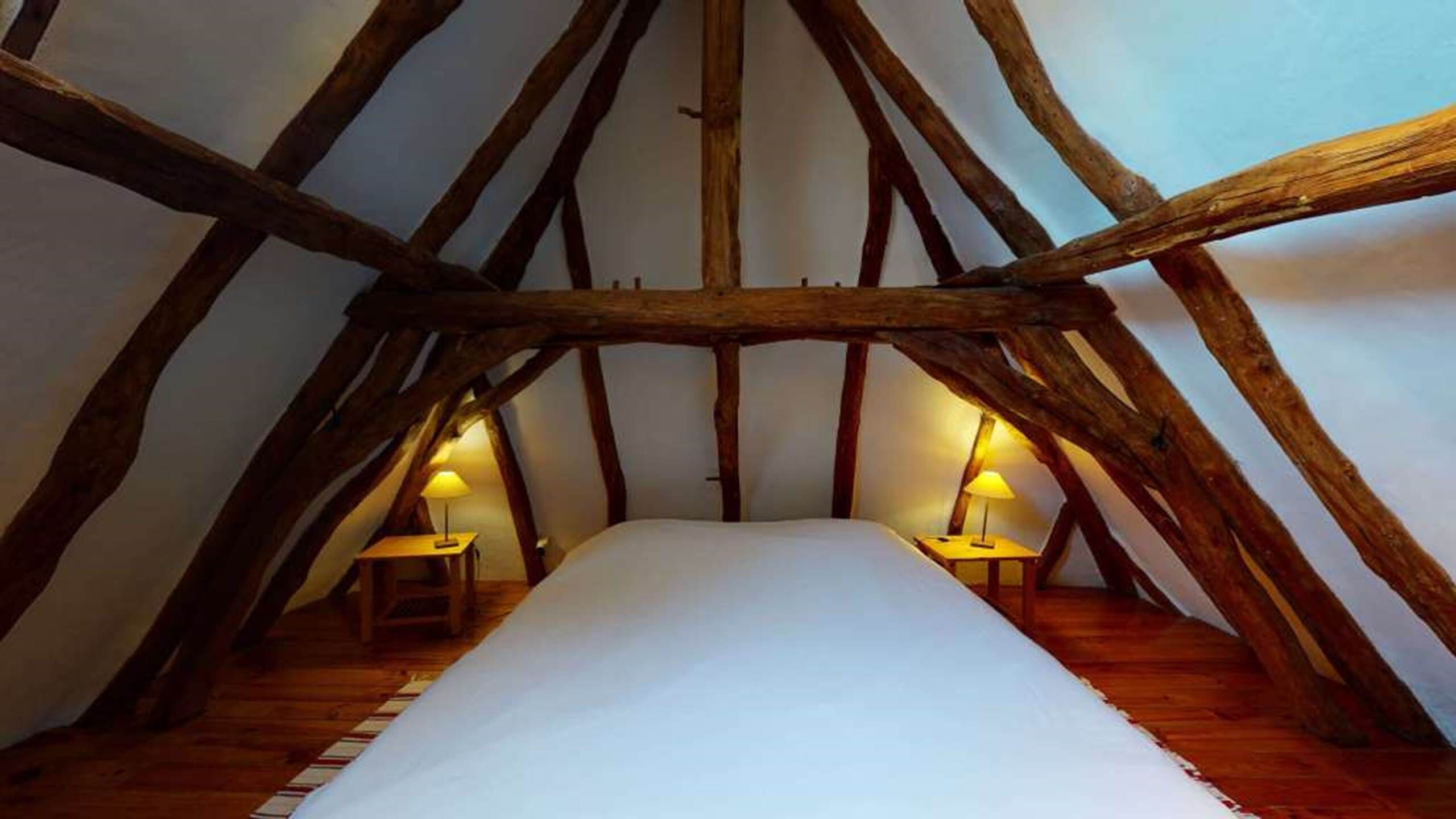 A high-ceilinged attic bedroom with exposed wooden beams, a central bed, and two bedside lamps.