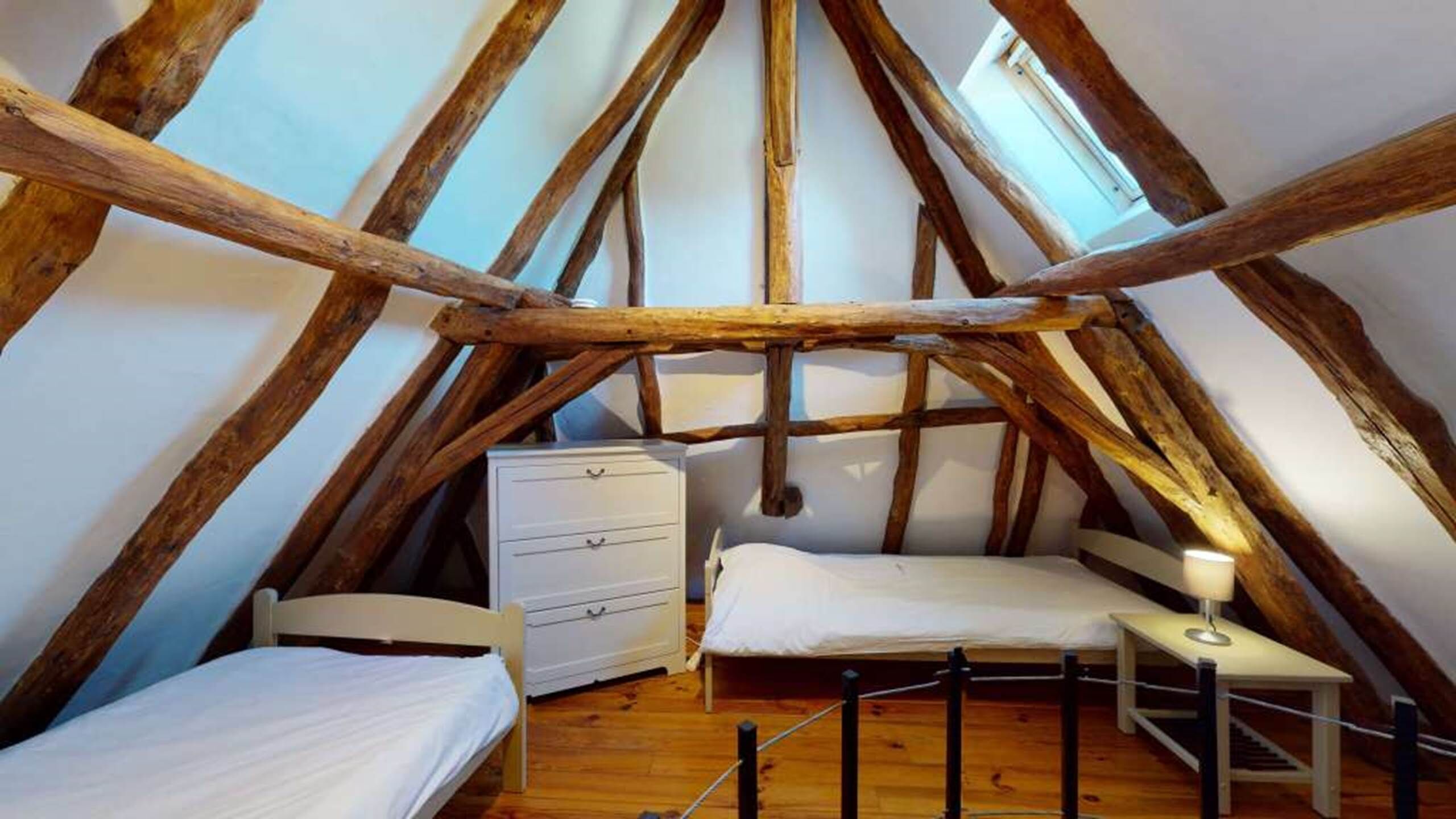A loft bedroom with a sloped ceiling, wooden beams, a bed, a dresser, and a nightstand.