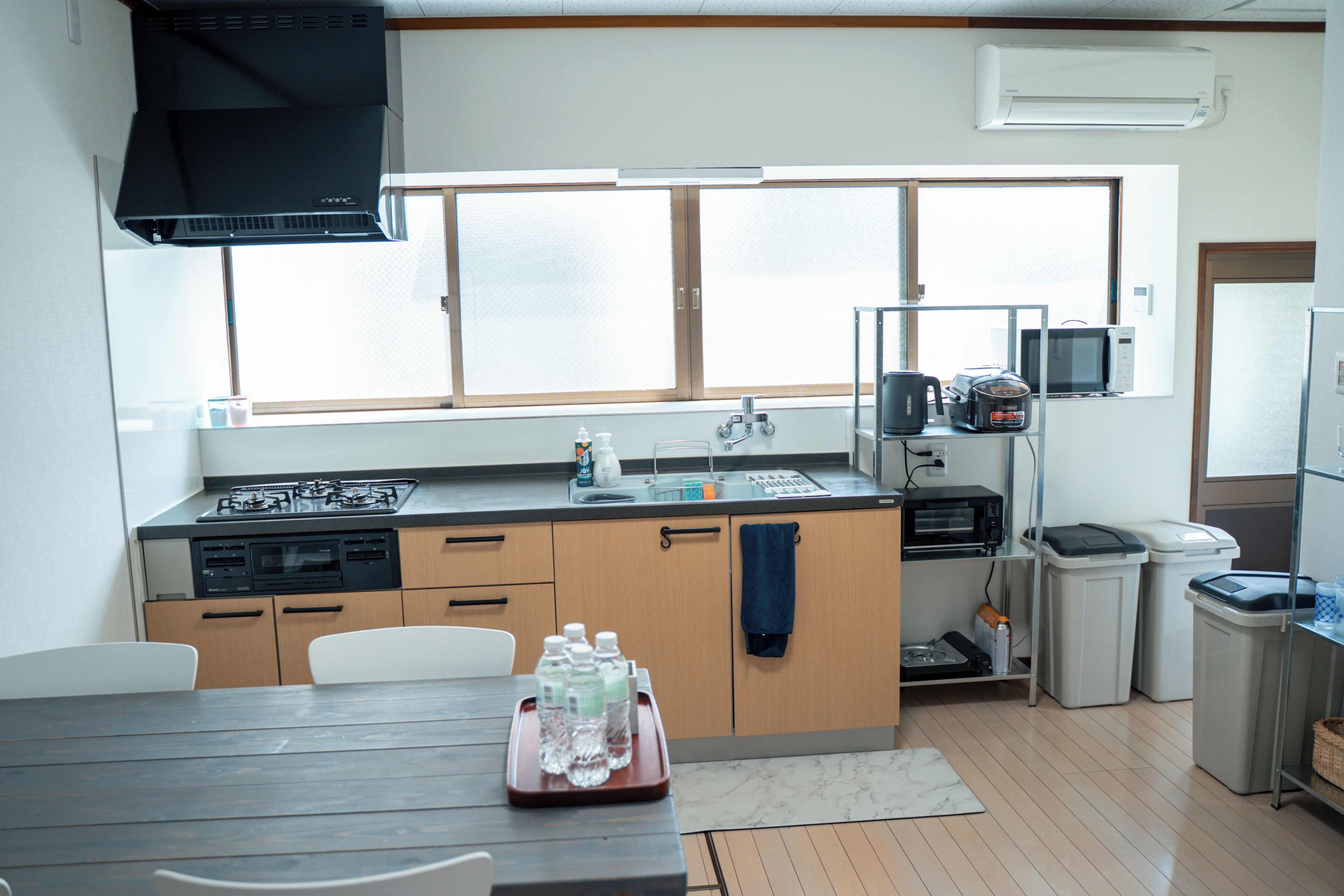 Una cocina con estufa, microondas y lavamanos, una mesa de comedor con sillas y una ventana con persianas.