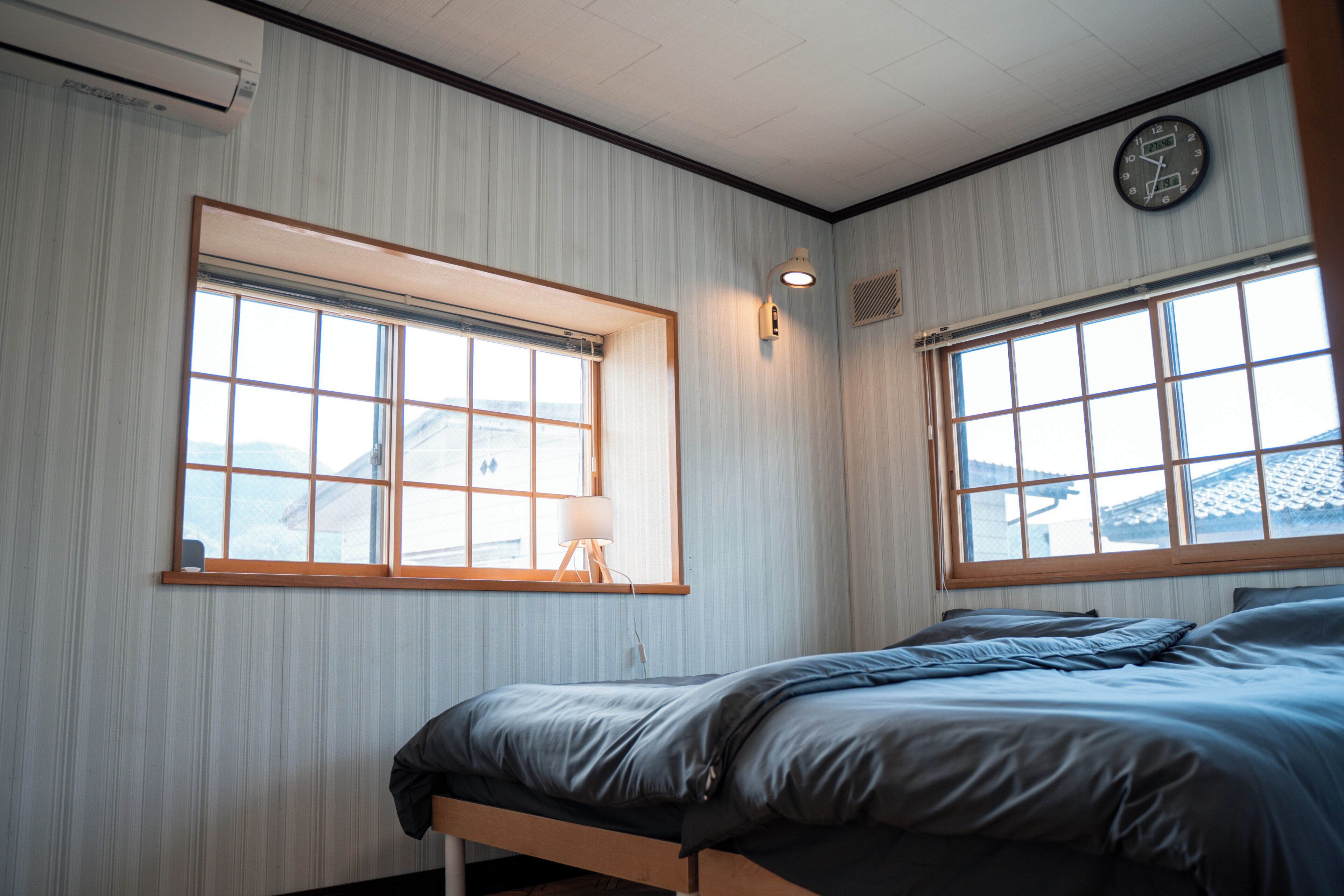 Une pièce avec deux fenêtres, un lit, une horloge murale et une lampe.