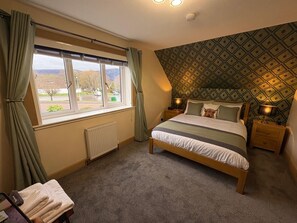 A bedroom with a bed, a radiator, a window with curtains, a patterned wall, and a nightstand with a lamp. - Kildonan House Bed and Breakfast (Fort William)