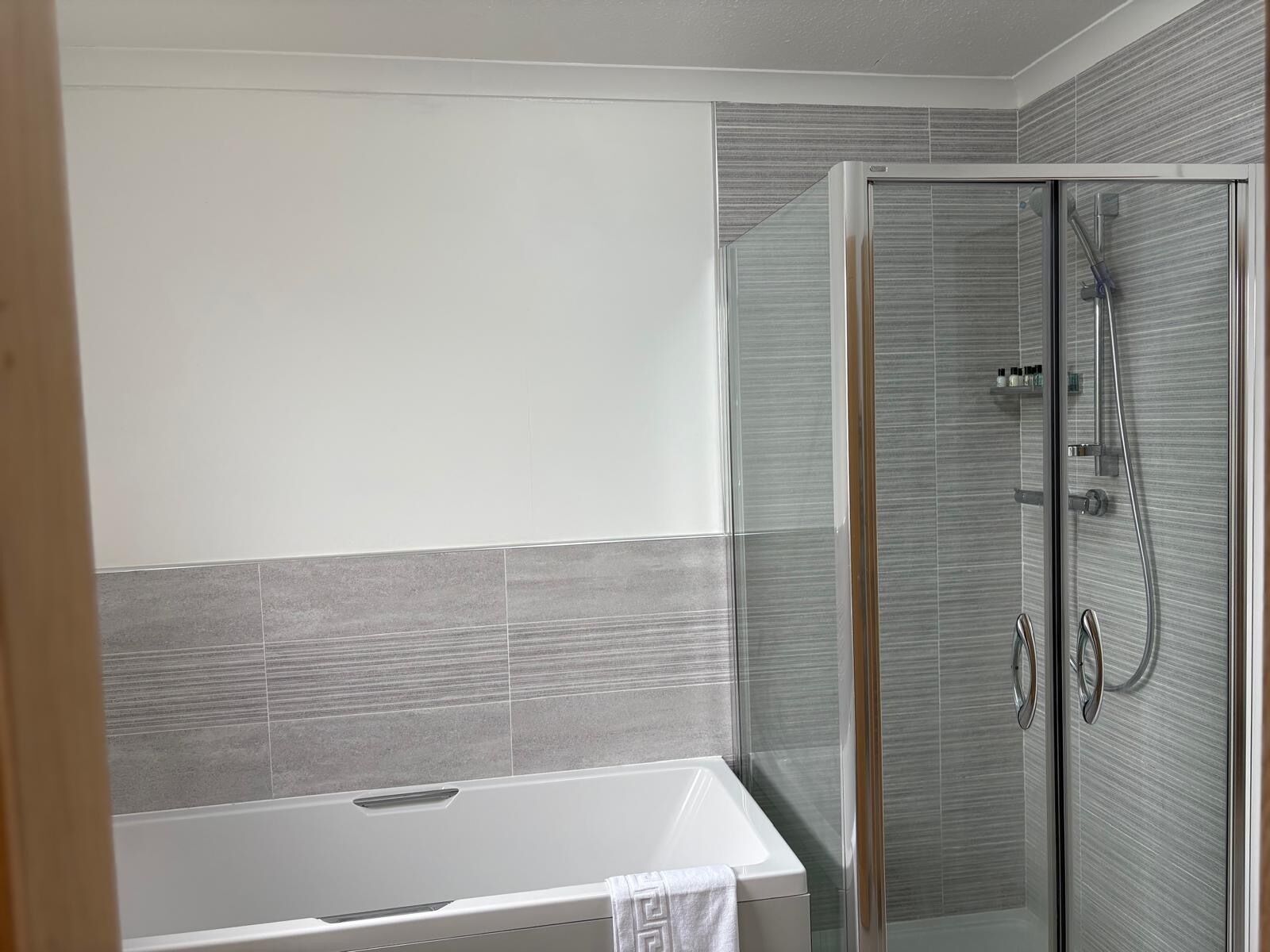 A modern bathroom with a glass-enclosed shower, a white bathtub, and grey tiled walls.