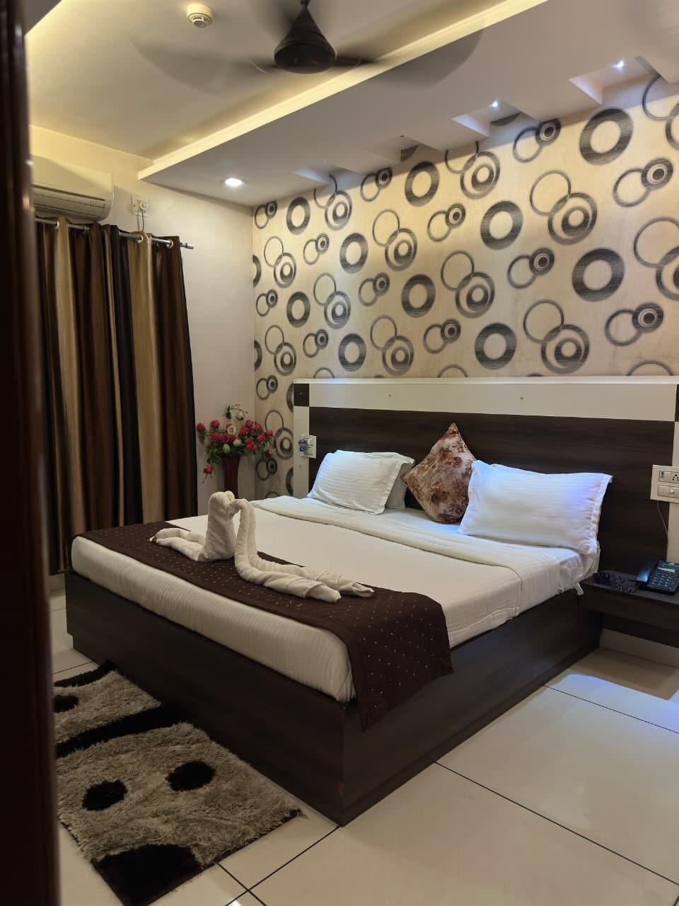 A modern hotel room with a large bed, a nightstand, a wall-mounted air conditioner, and a patterned wall.