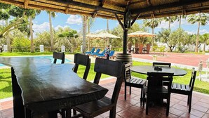 Una terrazza esterna con tavoli e sedie in legno, un tetto di paglia e una zona piscina sullo sfondo.