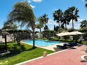 Una piscina con sillones de descanso y sombrillas, rodeada de palmeras y una estructura con techo de paja.