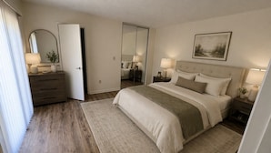 A bedroom with a bed, bedside tables, a dresser, a mirror, and a door leading to another room. - Beachside spacious 2-bedroom 2.5 bath townhome in Cedar Dunes. (New Smyrna Beach)