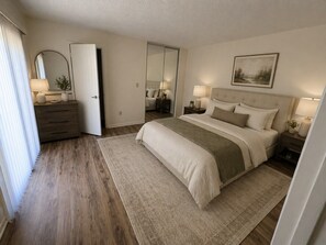 A bedroom with a bed, bedside tables, a dresser, a mirror, and a door leading to another room. - Beachside spacious 2-bedroom 2.5 bath townhome in Cedar Dunes. (New Smyrna Beach)