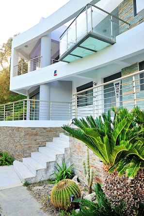 Ein modernes, zweistöckiges Haus mit einem Glasbalkon, einer Steintreppe und einer Vielfalt an Kakteen und Farne im Garten.