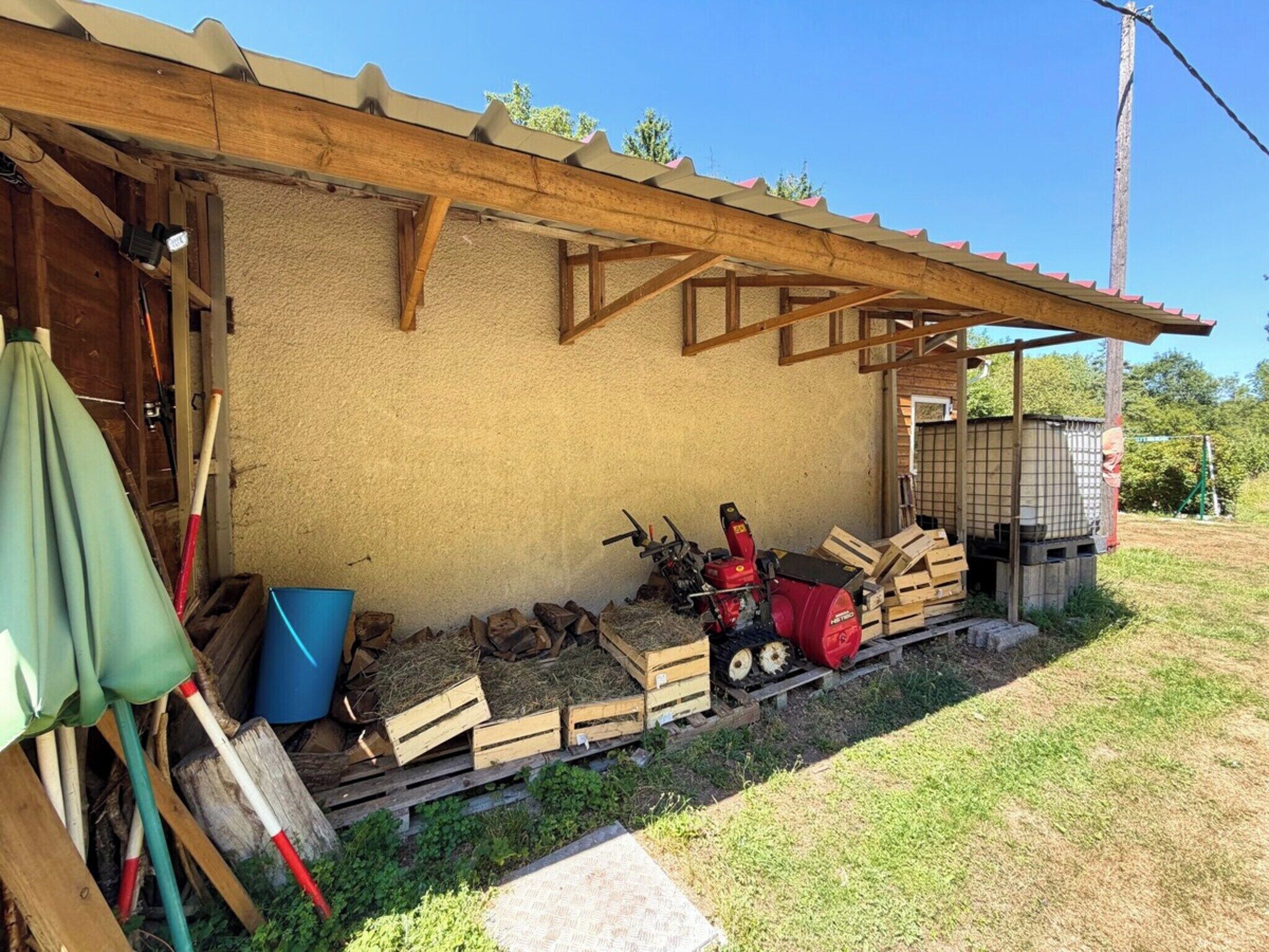 Une structure en bois surmontée d’un toit en tôle ondulée, une pile de bois de chauffage, une tondeuse à gazon rouge et un seau bleu.