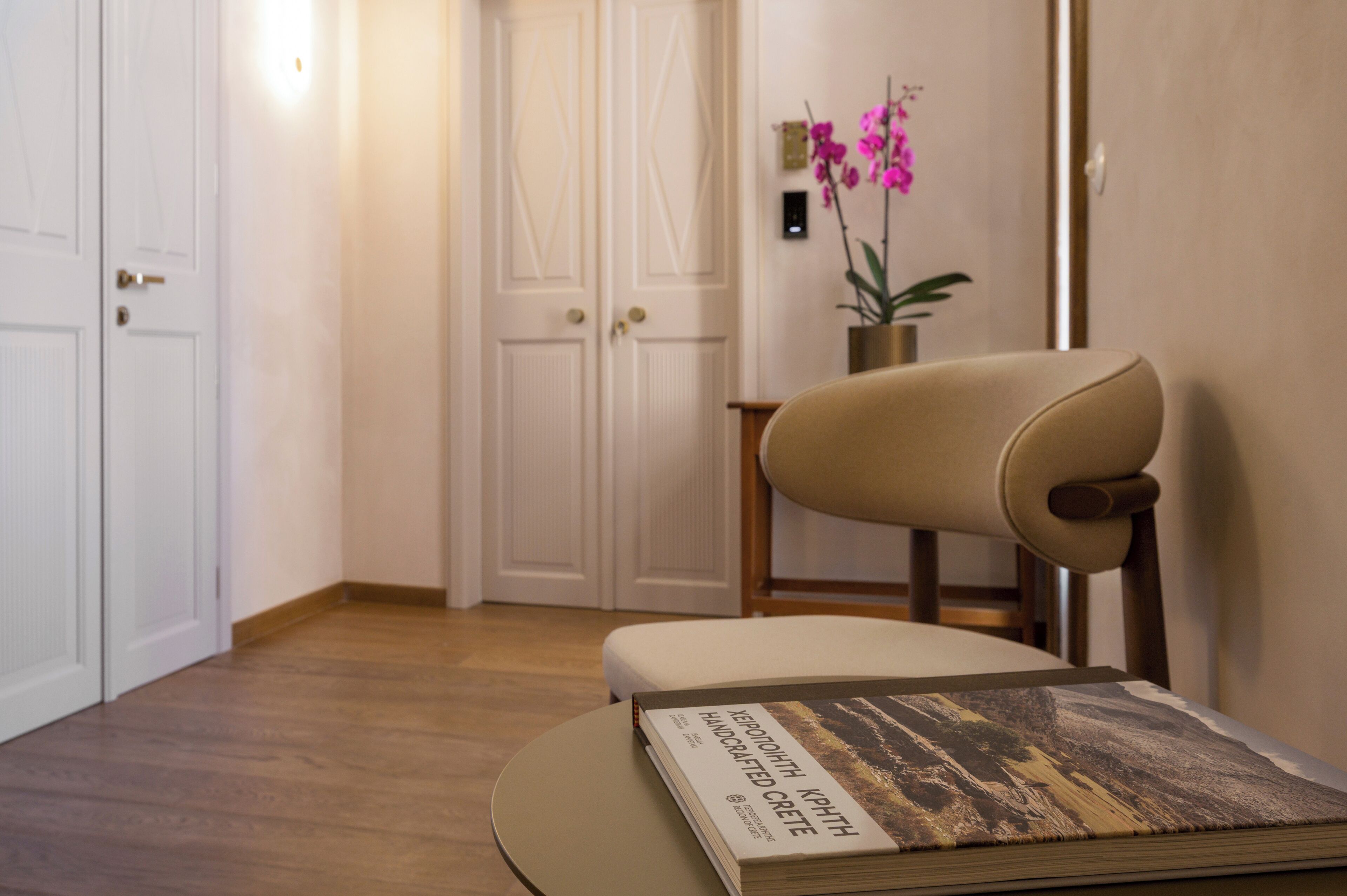 A room with a wooden floor, a beige chair, a small table with a book, and a door with a light switch.