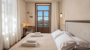 A hotel room with a bed, a desk, a chair, a radiator, a window with a view, and a door. - Palazzo Del Rettore  (Chania)
