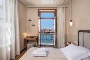 Quarto de hotel com uma cama, escrivaninha, cadeira, radiador, janela com vista para edifícios e varanda com guarda-corrimão.