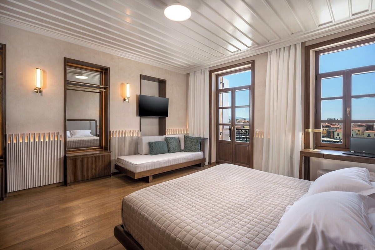 Quarto de hotel com uma cama grande, um sofá, uma televisão, uma janela com cortinas e vista para prédios.