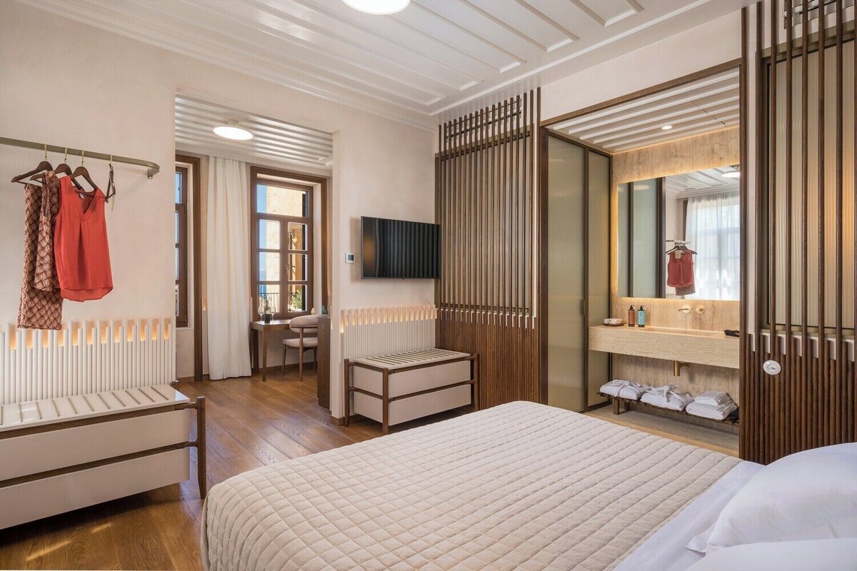 Quarto de hotel com uma cama, uma escrivaninha, uma cadeira, um radiador, um espelho e uma televisão.
