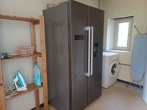 A compact kitchen with a stainless steel refrigerator, a washing machine, a wooden shelf with laundry items, and a window providing natural light. - Gîte le Bergerayre 6 people Swimming pool/Campagne 4-star accommodation (ST GRIEDE)
