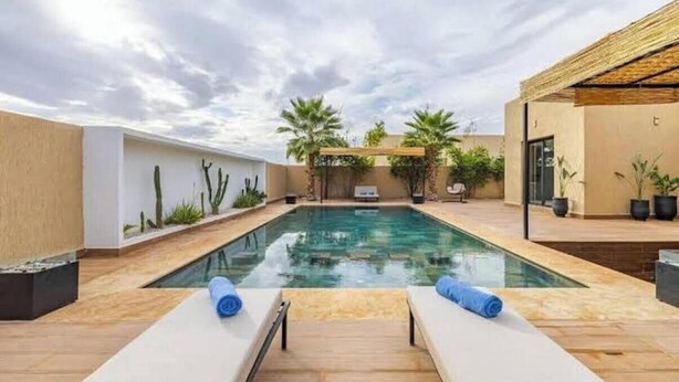 Un área de piscina moderna al aire libre con sillones de descanso, una pérgola y palmeras.