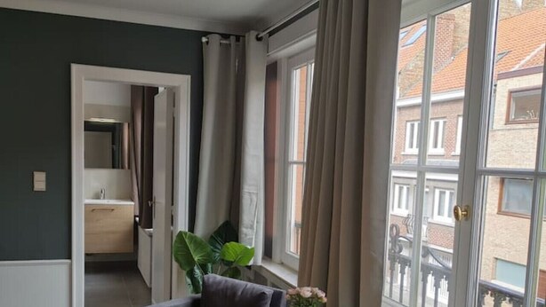 A modern living room with a sofa, armchair, and small table with a plant. A doorway leads to a kitchen area. - Spacious Quadruple Room w. Private Bathroom (Brugge)