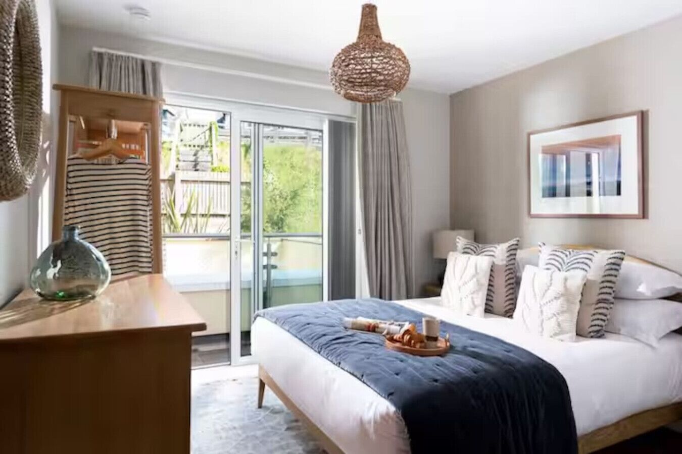 Ein Schlafzimmer mit einem großen Bett, einer hölzernen Kommode, einer Pendelleuchte und Blick ins Freie durch eine Glastür.
