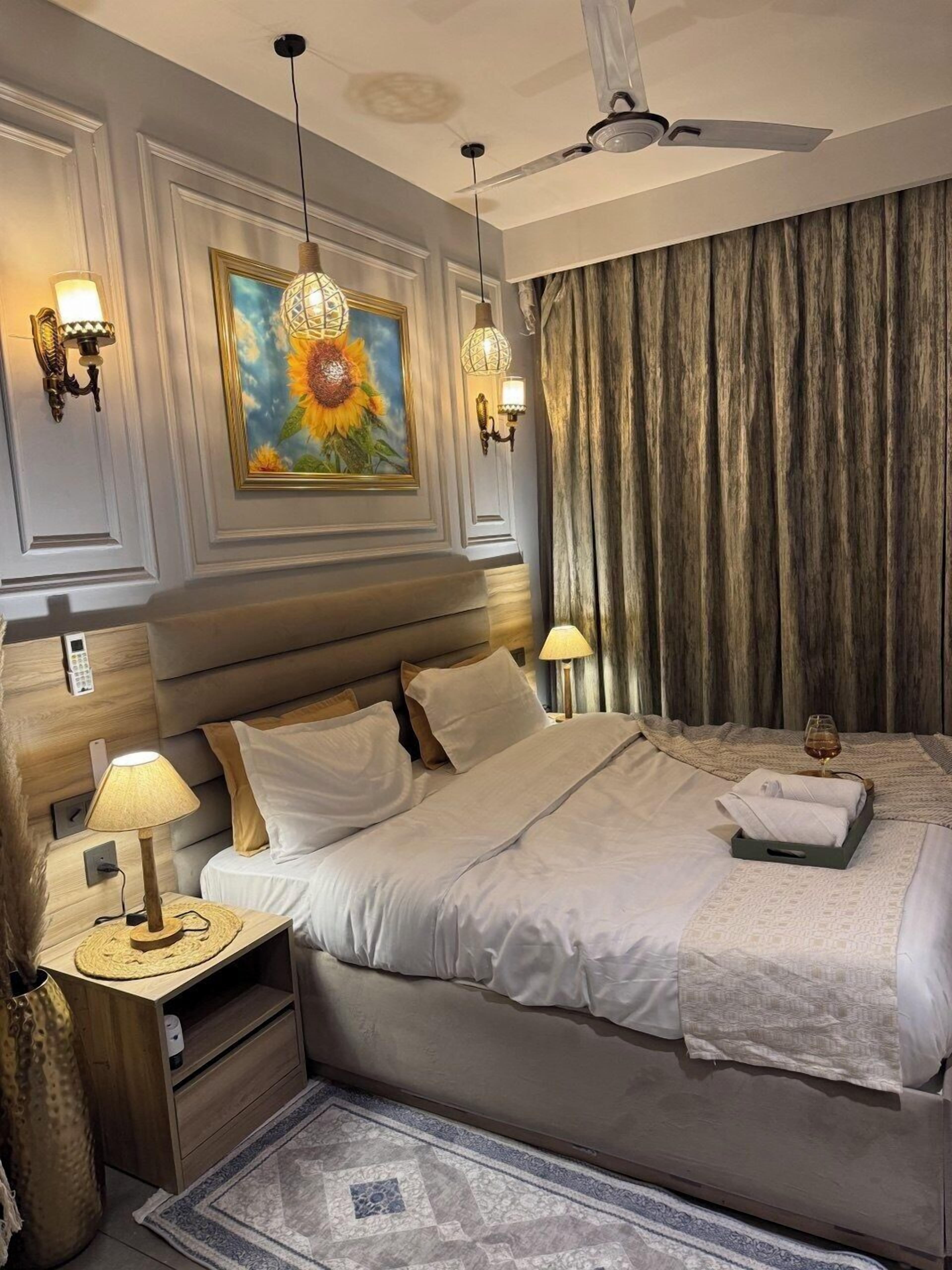 Una habitación de hotel con cama, lámparas de noche, un cuadro, ventilador de techo y ventana con cortinas.