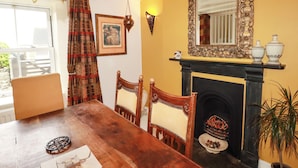 A dining room with a wooden table, chairs, a fireplace, a mirror, and a window with curtains. - Trelake Cottage (Tintagel)