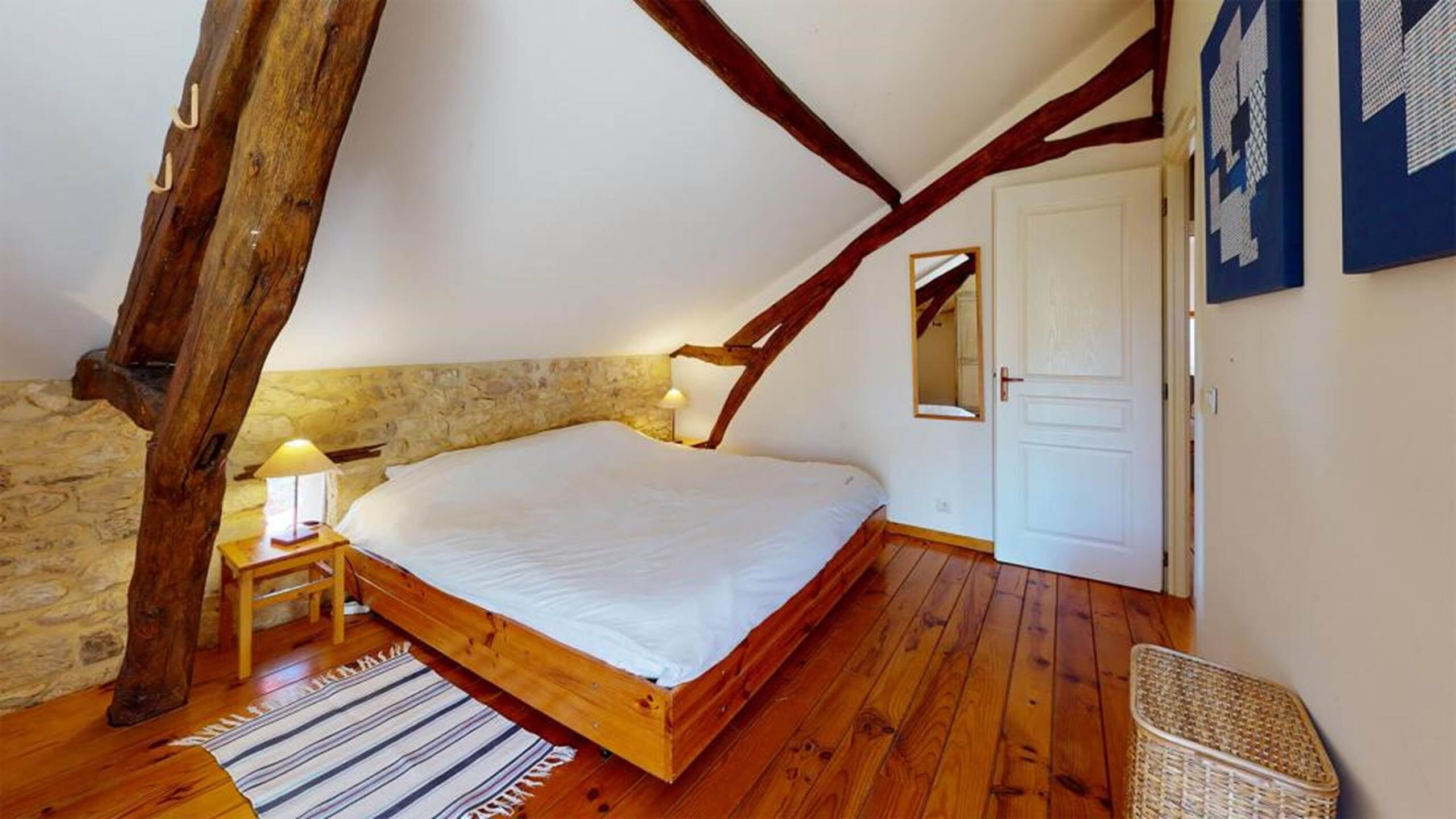 Une chambre avec un lit en bois, un mur en pierre, des poutres apparentes, une petite table de chevet avec une lampe et un panier en osier.