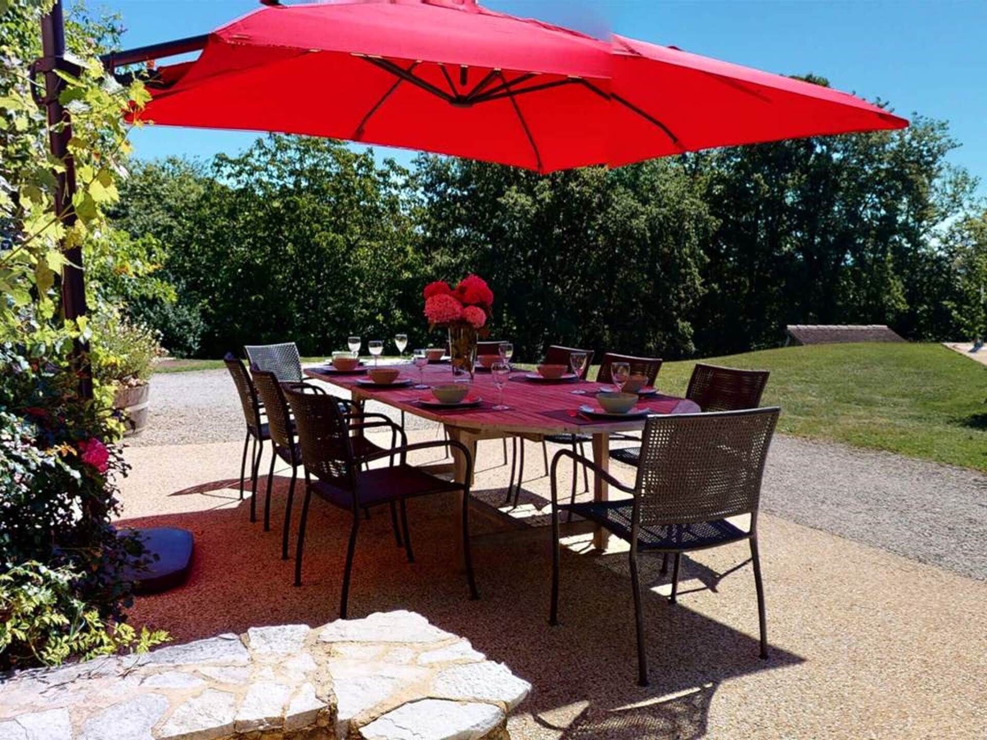 An outdoor patio with a table set for dining, surrounded by chairs, under a red umbrella.