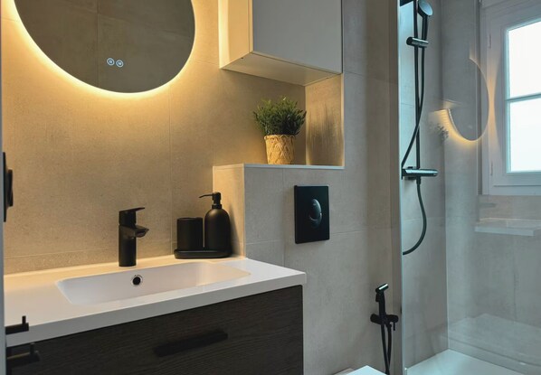 A modern bathroom with a white sink, black fixtures, a round mirror, a potted plant, and a glass shower enclosure. - Le vert Boulogne  (Boulogne-Billancourt)