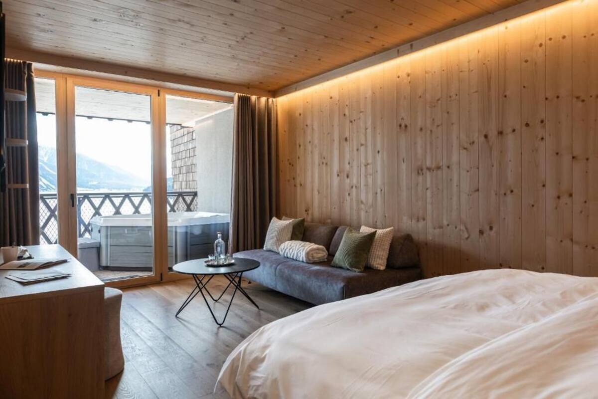 Habitación de hotel con techo de madera, cama, sofá, una pequeña mesa redonda y balcón con vistas a montañas.