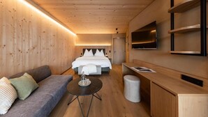A modern hotel room with a wooden ceiling, a bed with white bedding, a sofa, a small round table, and a TV mounted on the wall. - Das Gerstl Family Retreat (Curon Venosta)