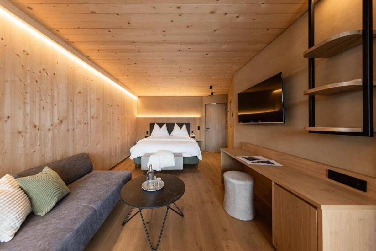 Una habitación de hotel moderna con techo de madera, una cama con ropa blanca, un sofá, una mesita redonda y un televisor montado en la pared.
