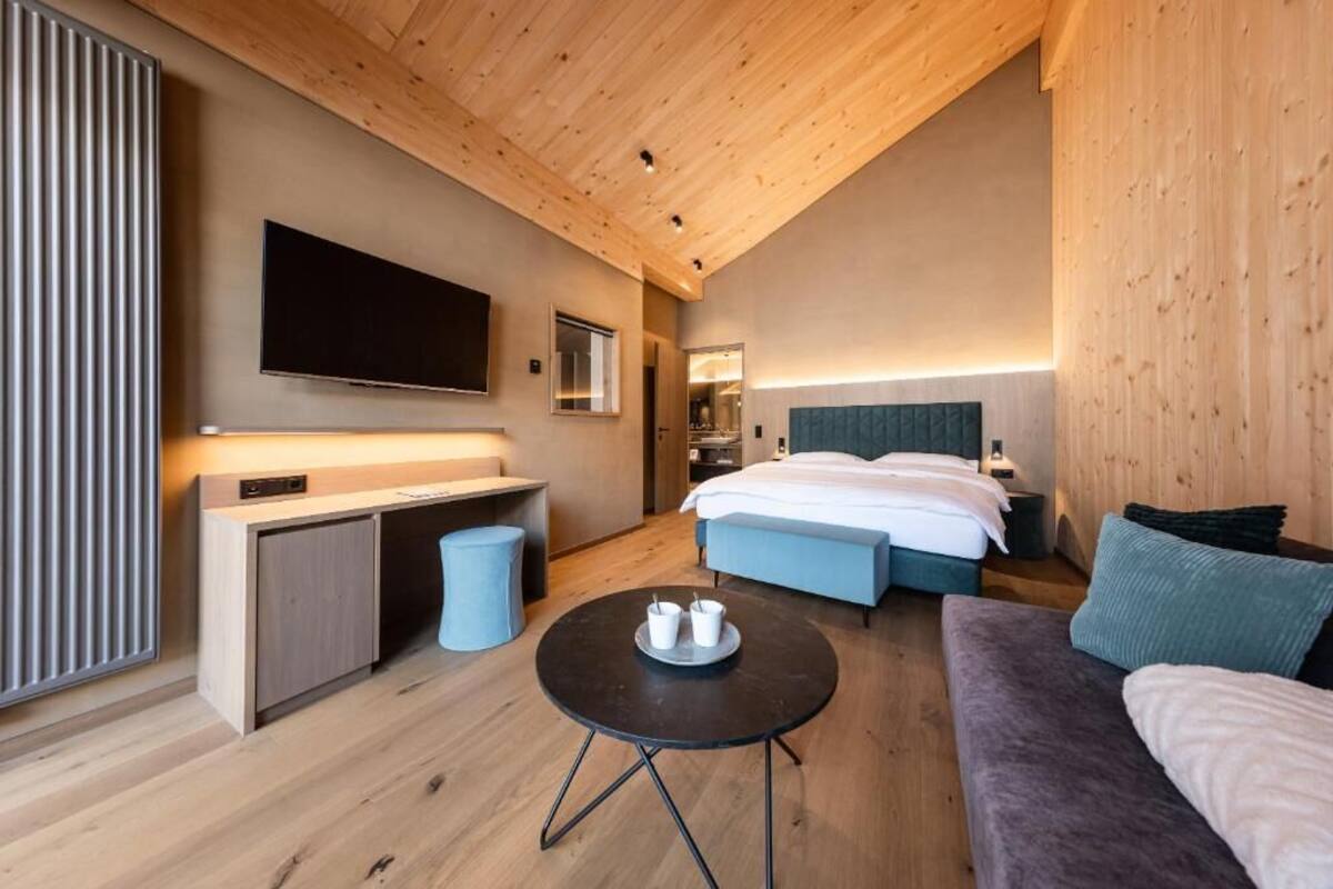 Una habitación de hotel moderna con techo de madera, televisor de pantalla plana, cama con ropa blanca, sofá y una pequeña mesa redonda con tazas.