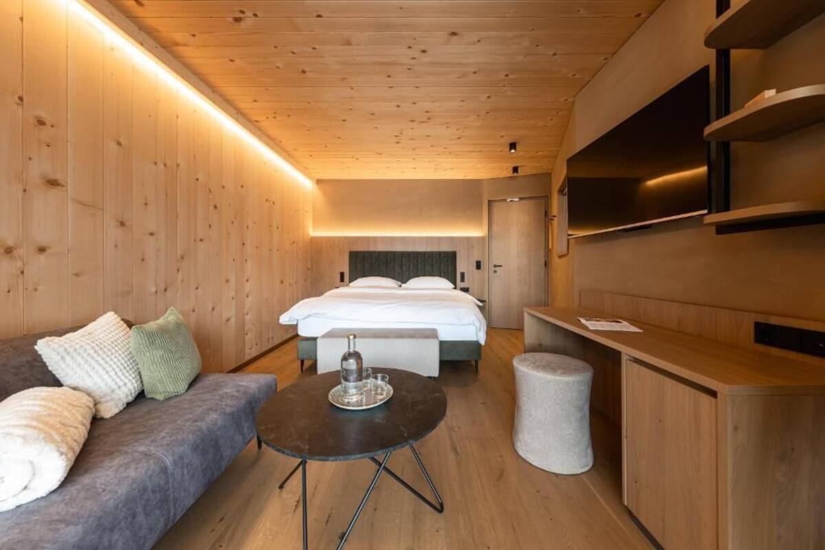 Una habitación de hotel moderna con techo de madera, una cama con sábanas blancas, un sofá gris, una pequeña mesa redonda con una bandeja y un armario de madera.